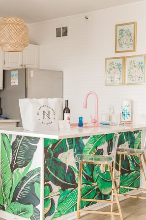 A kitchen with a white refrigerator, a sink with a pink faucet, and a bar area with chairs. There are framed botanical prints on the wall.