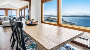 Un vaste espace dédié à la salle à manger, avec une longue table en bois, des chaises noires et une vue sur la mer grâce à de grandes fenêtres.