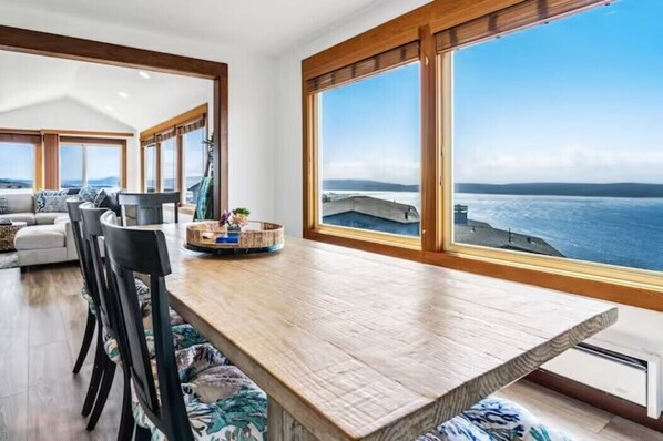 Un comedor amplio con una larga mesa de madera, sillas negras y vistas al mar a través de grandes ventanales.