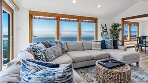 Un salon doté d’un grand canapé d’angle, d’une table basse ronde en bois et offrant une vue sur la mer grâce à de grandes fenêtres.