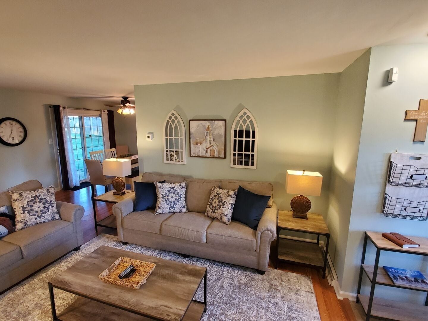A living room with a sofa, a coffee table, and a lamp. There is a window with curtains, a wall clock, and a cross on the wall.
