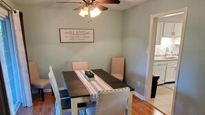 Une salle à manger avec une table en bois, des chaises, un ventilateur de plafond et une cuisine visible à travers une porte ouverte.