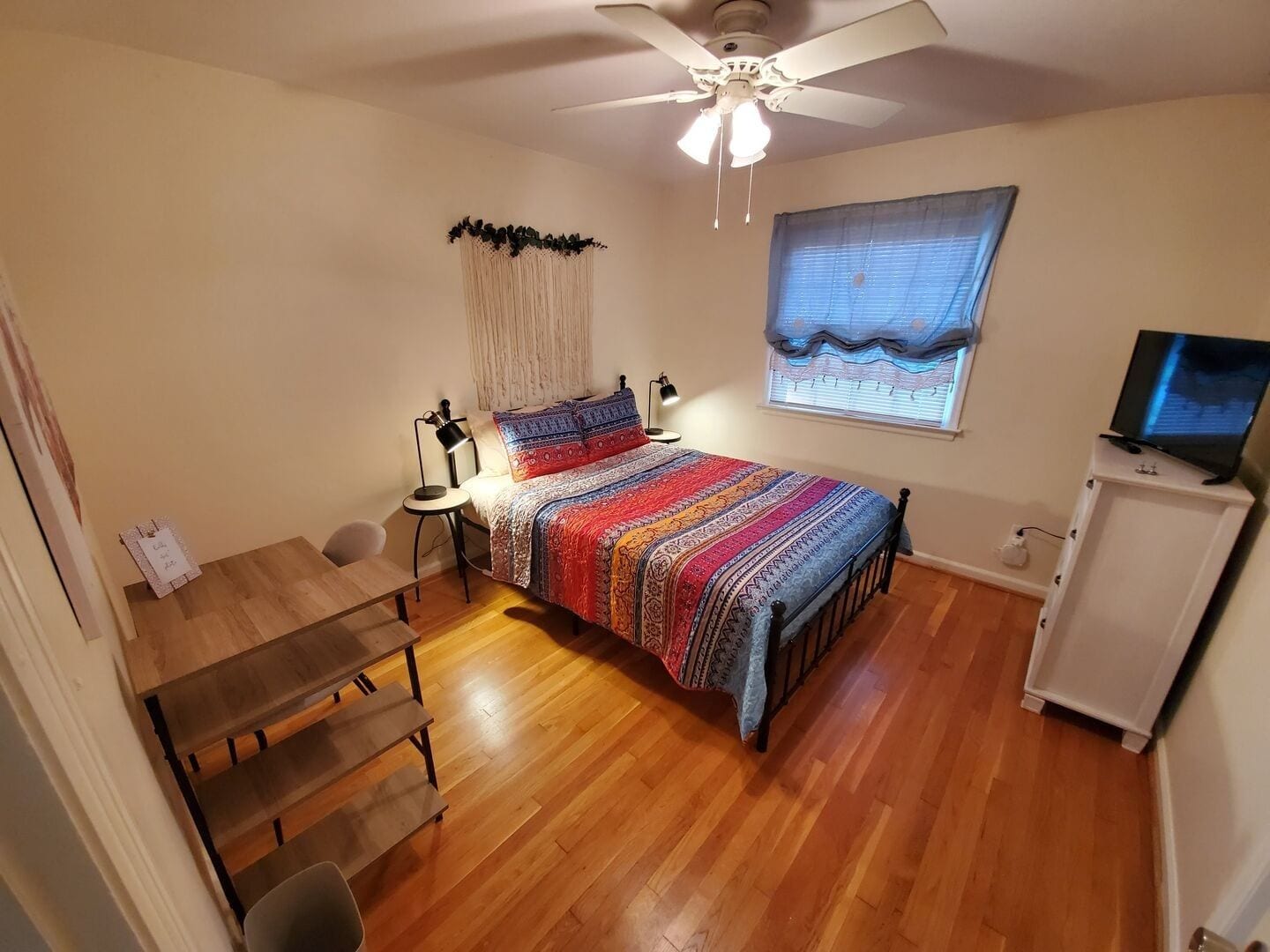 A bedroom with a bed, a desk, a chair, a ceiling fan, a window with blinds, and a television on a cabinet.