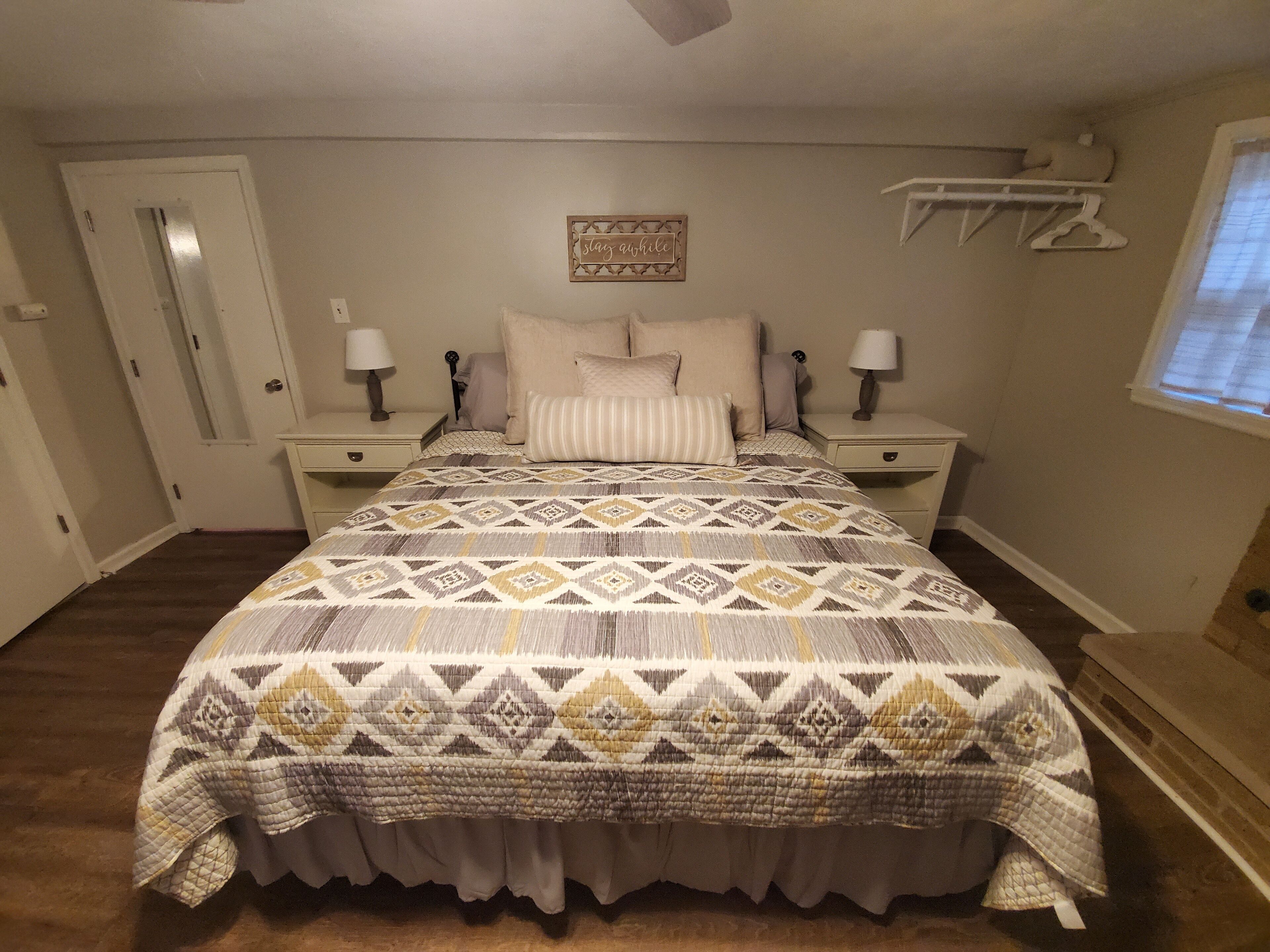 A bedroom with a patterned bedspread, two bedside tables with lamps, a door, and a wall-mounted shelf with a decorative item.