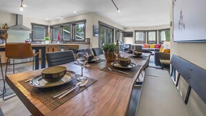 Une cuisine moderne avec des meubles de couleur sombre, un coin repas avec une table en bois dressée pour quatre personnes, et un salon avec un canapé et des coussins colorés.