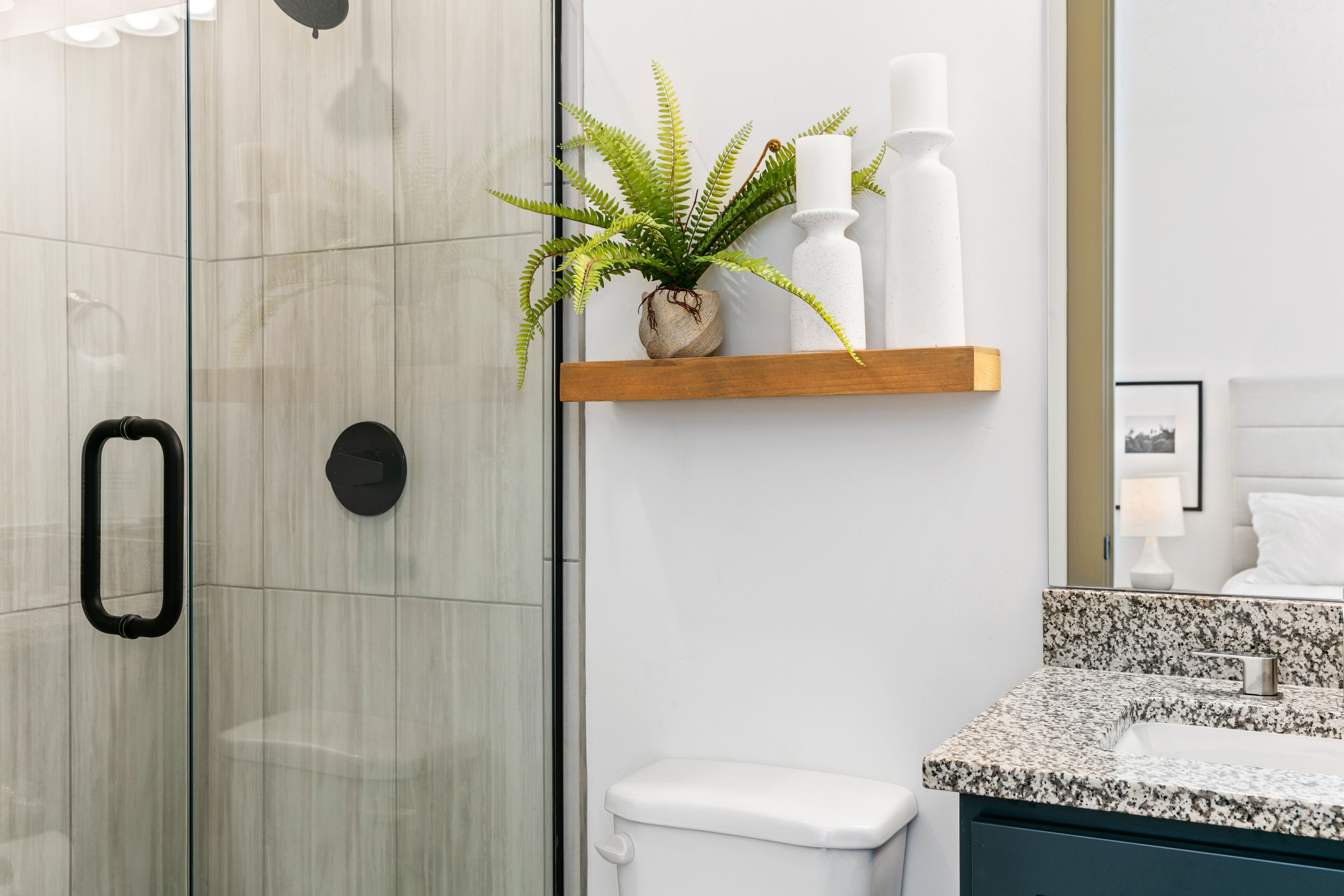 A modern bathroom with a glass shower enclosure, a white toilet, a granite countertop sink, and a shelf with a potted plant and candles.