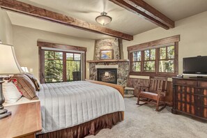 A bedroom with a large bed, a stone fireplace, wooden beams, a television, and a view of trees. - Escape to this stunning private home in Breckenridge! (Breckenridge)