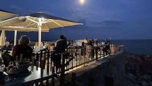 An outdoor dining area with tables and chairs, a railing, and a view of the sea.