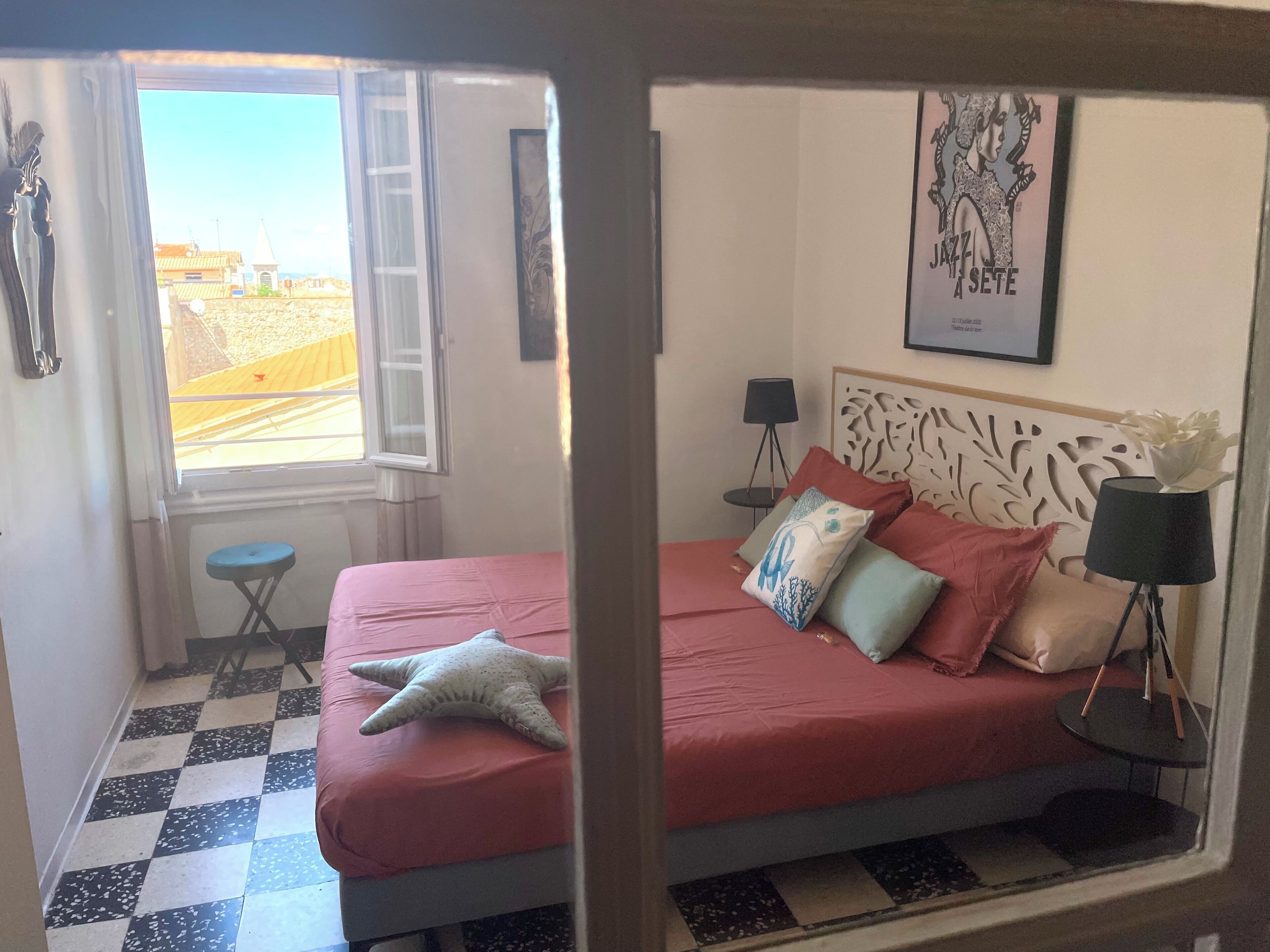 A bedroom with a checkered floor, a bed with pink bedding, a starfish decoration, a small stool, and a window with a view of buildings.