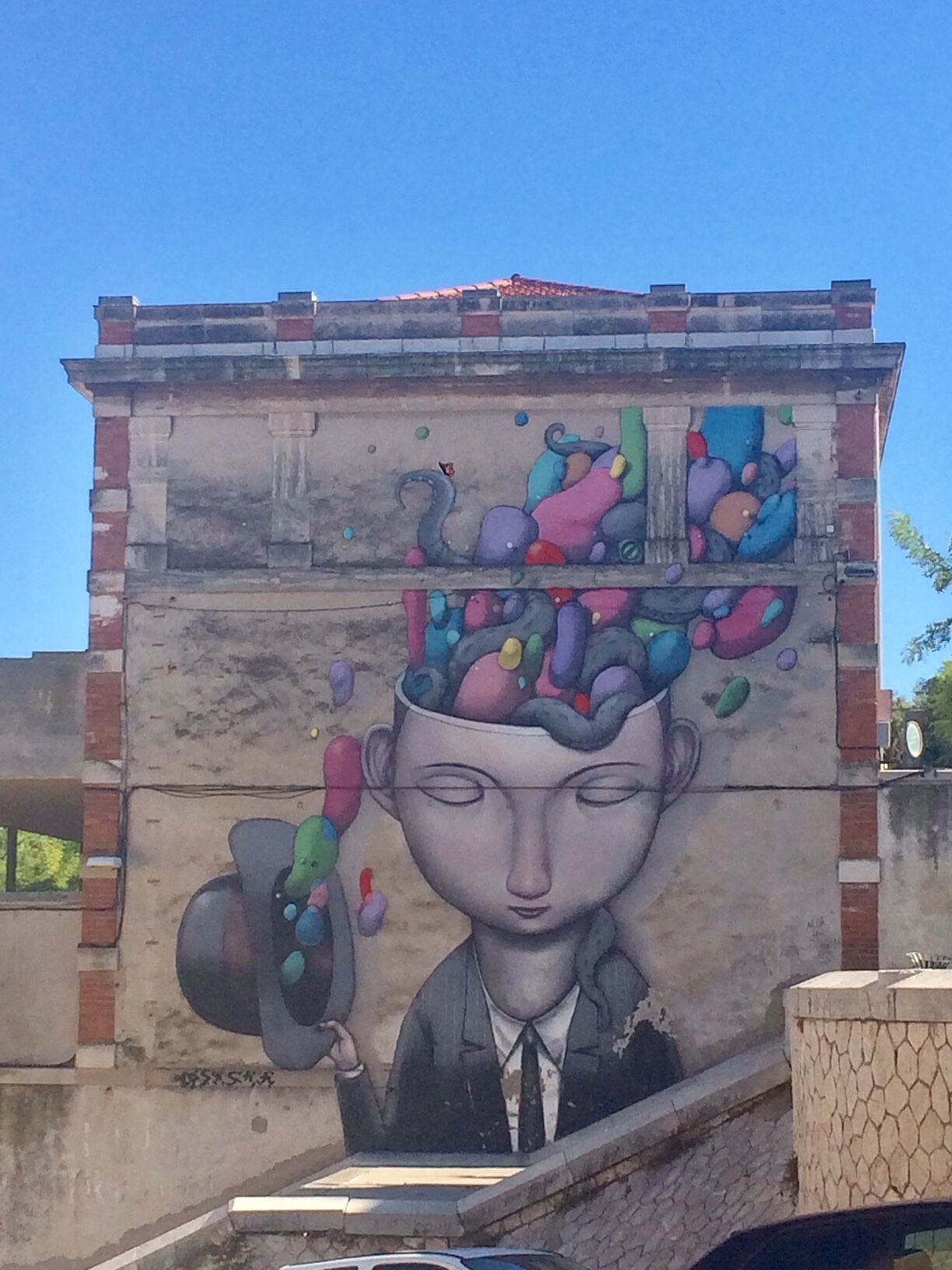 Une fresque orne un bâtiment : on y voit une personne tenant un chapeau, et des formes colorées au-dessus de sa tête.