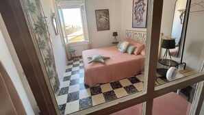 A bedroom with a checkered floor, a bed with pink bedding, a small table with a lamp, and a mirror on the wall.