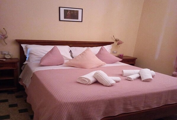 A neatly made bed with pink bedding and pillows, a framed picture on the wall, and a small clock on the nightstand. - Near the center (Lucca)