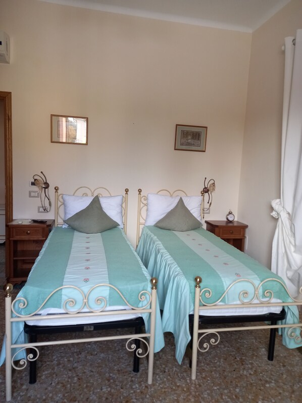 Una habitación con dos camas, cada una con estructura metálica y cubrecama azul claro. Hay dos mesitas de noche con relojes y un cajón pequeño. Las paredes son lisas, y hay un cuadro enmarcado en la pared.