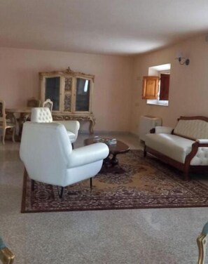 A living room with a white sofa, a white armchair, a coffee table, and a patterned rug. There is a wooden cabinet and a window with a wooden shutter. - Near the center (Lucca)