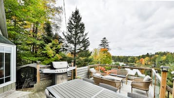 Une terrasse en bois, meublée pour l’extérieur, avec un barbecue et une vue sur un lac entouré d’arbres.