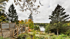 A wooden deck with a staircase leading to a covered area, surrounded by a garden with various plants and a view of a lake.