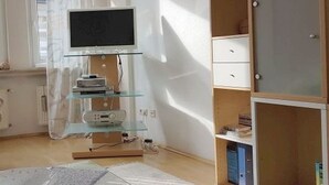 Una habitación con televisor sobre una repisa de vidrio, un mueble de madera con cajones y una ventana con cortinas.