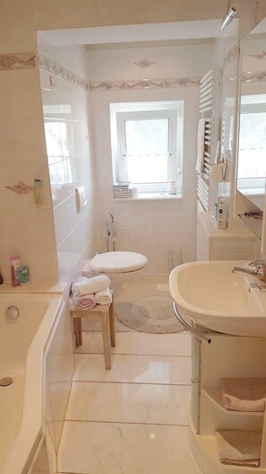 Un baño con bañera, inodoro y lavabo. Cuenta con una ventana que proporciona luz natural y un perchero para toallas.