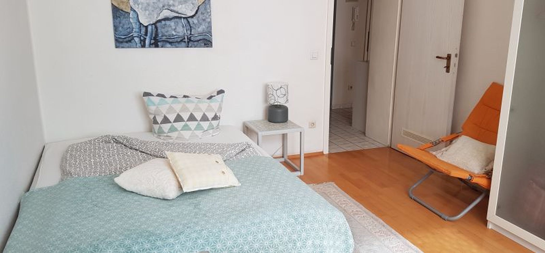 Ein Schlafzimmer mit einem Bett, Kissen, einem kleinen Tisch mit Lampe, einem Sessel und einer Tür zu einem anderen Raum.