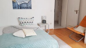 Un dormitorio con cama, almohadas, una mesita de noche con lámpara, una silla y una puerta que da a otra habitación.