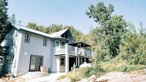 A two-story house with a balcony, surrounded by trees and a clear sky. - Private beach, boat slip, hot tub, pool table, fenced yard, BBQ, fire pit (Bass Lake)