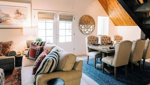 A cozy living room with a sofa, a dining table with chairs, and a large window with blinds. - Private beach, boat slip, hot tub, pool table, fenced yard, BBQ, fire pit (Bass Lake)