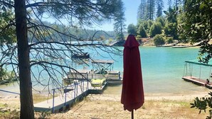 A picnic table with a red umbrella, surrounded by a natural setting with trees and a lake. - Private beach, boat slip, hot tub, pool table, fenced yard, BBQ, fire pit (Bass Lake)