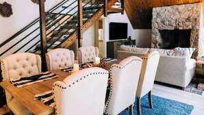 A dining area with a wooden table and white upholstered chairs, a stone fireplace, and a staircase with a wooden railing. - Private beach, boat slip, hot tub, pool table, fenced yard, BBQ, fire pit (Bass Lake)