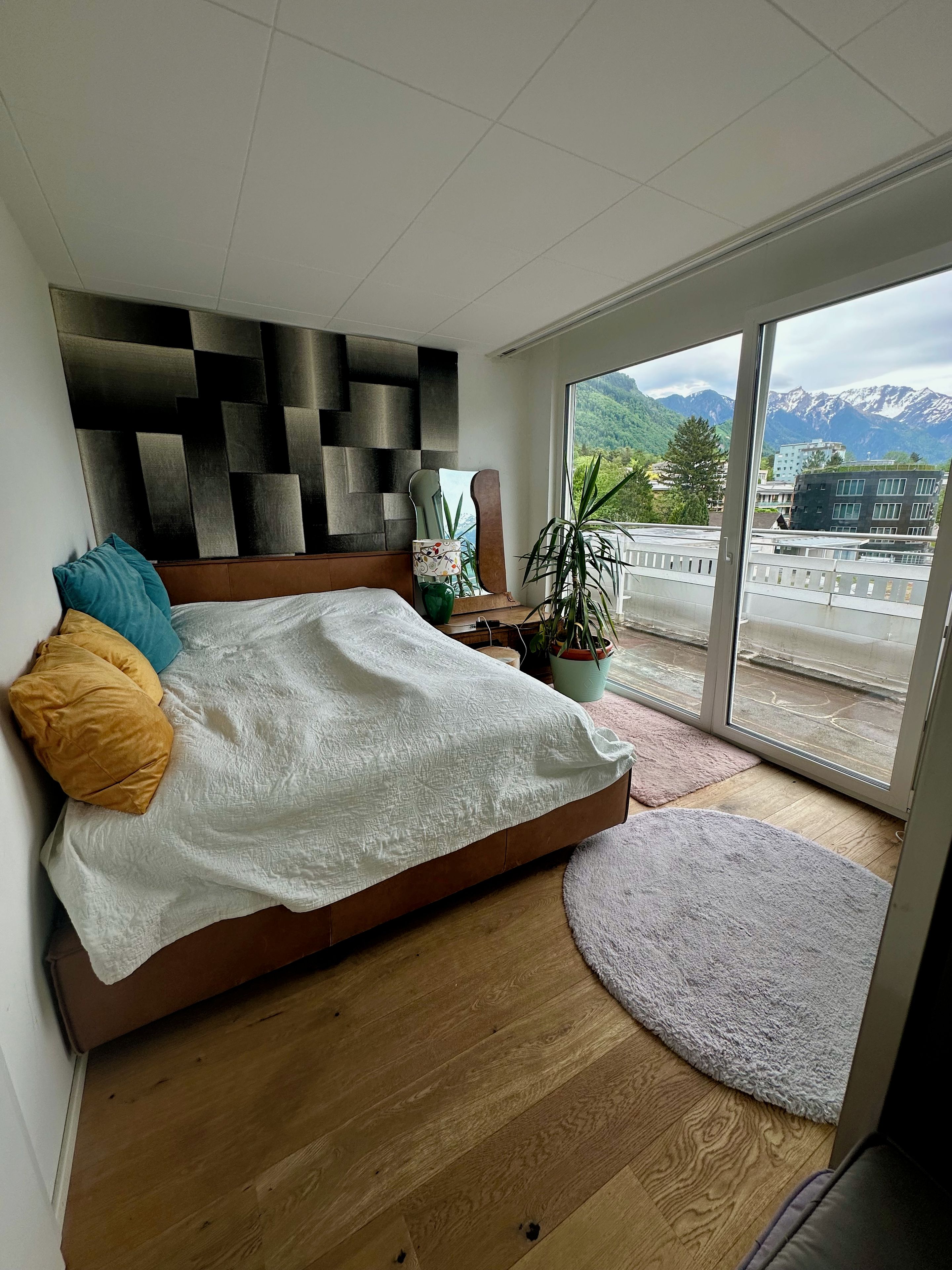 Une chambre à coucher avec un lit, une tête de lit ornée d’un motif géométrique, une plante en pot et une vue sur les montagnes par la fenêtre.