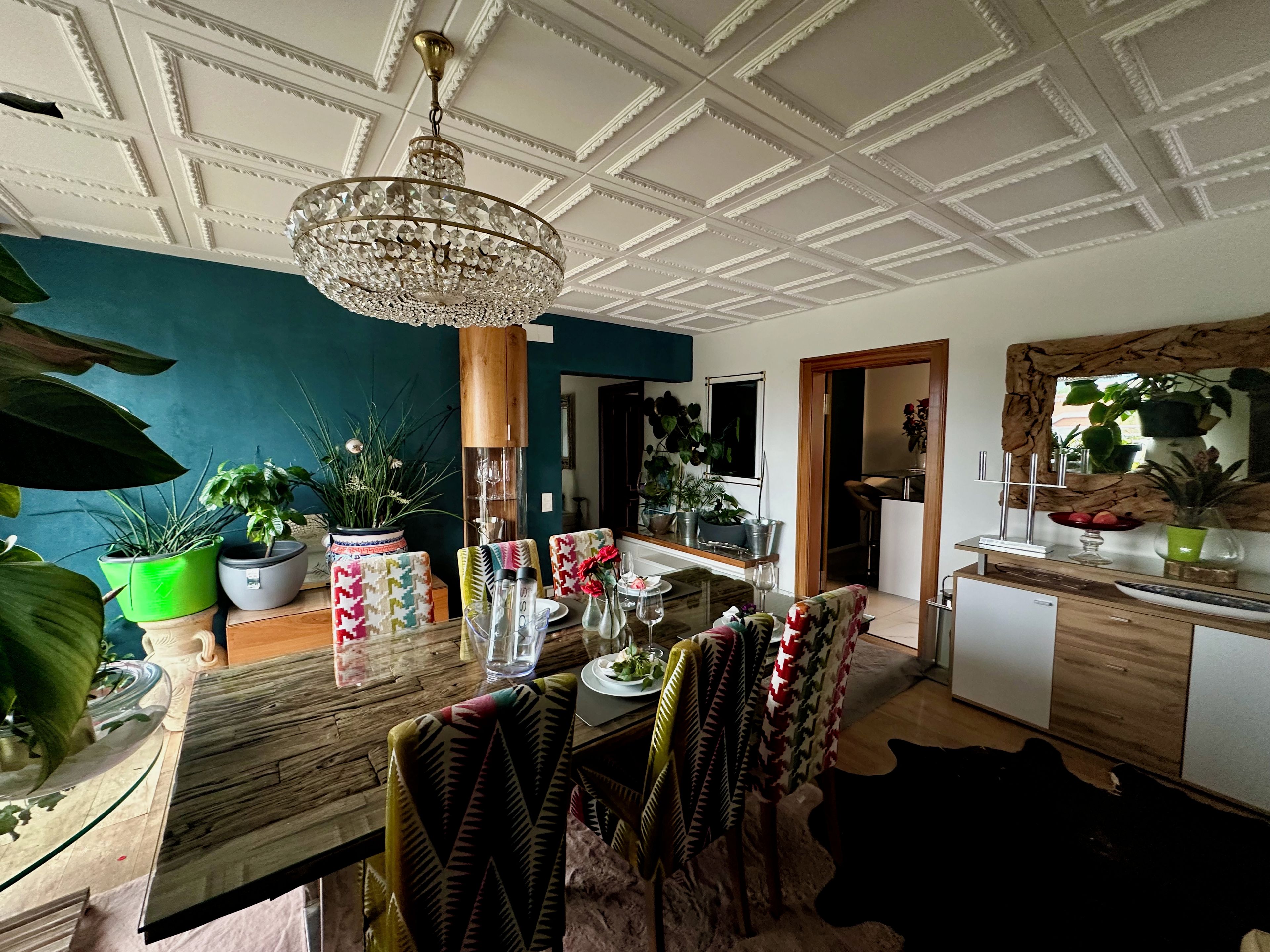 Un comedor con mesa de madera, sillas y una araña. Hay plantas en macetas y un espejo en las paredes.