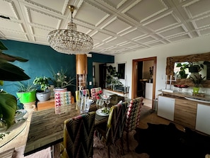 A dining area with a wooden table, chairs, and a chandelier. There are potted plants and a mirror on the walls.