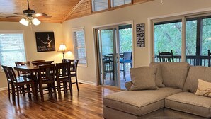 A spacious living room with a wooden floor, a sectional sofa, a dining area with a table and chairs, and large windows offering views of the outdoors. - Cozy Lake Hartwell Cabin - Beautiful dock views! Bring your boat and your pet! (Lavonia)
