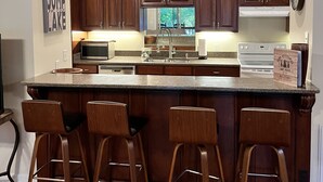 A kitchen with wooden cabinets, a large island with bar stools, a microwave, and a clock on the wall. - Cozy Lake Hartwell Cabin - Beautiful dock views! Bring your boat and your pet! (Lavonia)