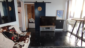 Un salon moderne avec un plancher noir, un canapé agrémenté de coussins à motifs, un meuble pour la télévision et un fauteuil placé près d’une fenêtre.