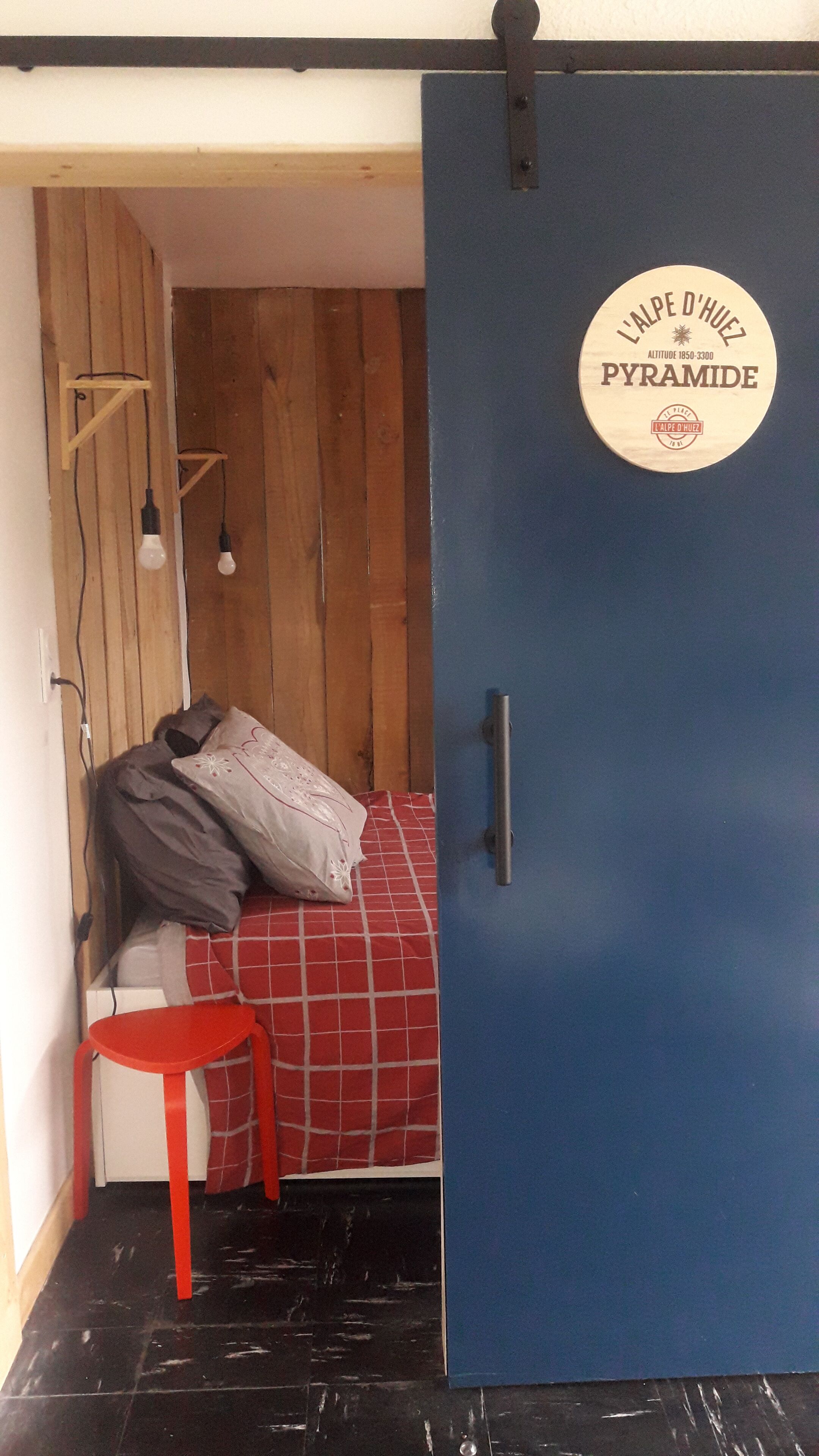 A room with a blue door, a red chair, a bed with a red and white checkered blanket, and a wooden wall.