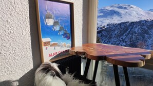 Une photo encadrée représentant un télésiège, avec l’inscription « ALPE D’HUEZ ISÈRE – FRANCE » sur un mur, une table en bois avec une chaise, et un chien qui regarde par la fenêtre.