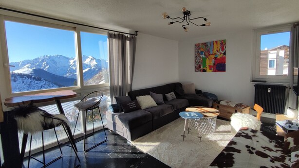 Un salon moderne avec un canapé noir, une petite table basse et une vue sur des montagnes enneigées, visible à travers de grandes fenêtres.