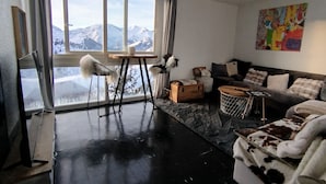 Un salon moderne doté d’une grande fenêtre offrant une vue sur les montagnes, d’un sol en marbre noir, d’un canapé agrémenté d’un plaid en peau de vache et d’un tableau abstrait aux couleurs vives accroché au mur.