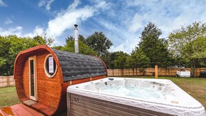 A hot tub and a wooden barrel-shaped structure with a chimney, set in a backyard with a wooden fence and trees.