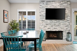 A dining area with a stone fireplace, a flat-screen TV mounted above it, and a large window with a view of the outdoors.