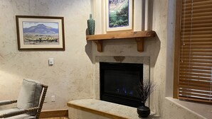 Ein Wohnzimmer mit Kamin, einem gerahmten Landschaftsbild, einer Vase und einem Sessel.