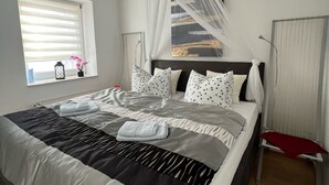 Un lit bien fait, recouvert d’une couvre-lit à rayures noires et blanches, surmonté d’un baldaquin et agrémenté d’un coussin à motifs bleus. On aperçoit une fenêtre avec des stores, une table de chevet avec une lampe et un tableau accroché au mur.