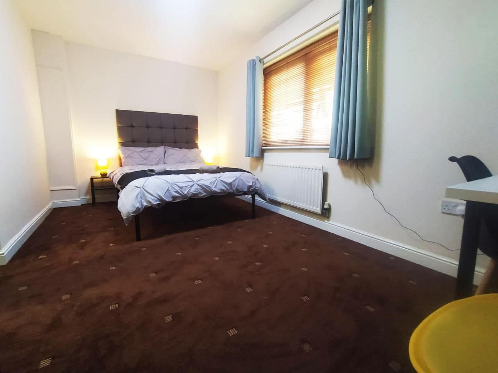 A bedroom with a bed, bedside lamps, a radiator, a window with curtains, and a desk with a chair.