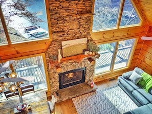 A cozy cabin interior with a stone fireplace, wooden walls, large windows, and a dining area.