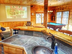 L’intérieur d’un chalet en bois est aménagé avec une table de billard, un évier en pierre et un écran de projection qui diffuse une sélection de jeux.