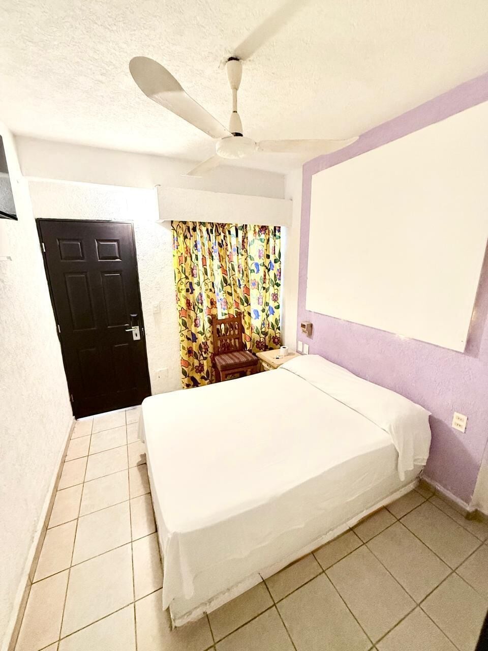 A room with a bed, a ceiling fan, a door, and a curtain with a floral pattern.