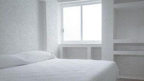 Une pièce blanche avec un lit, une fenêtre et des étagères intégrées.