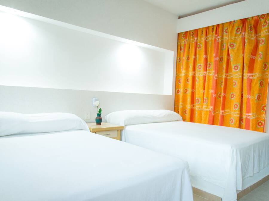 Quarto de hotel com duas camas de solteiro, mesa de cabeceira com um vaso, e cortina laranja vibrante.
