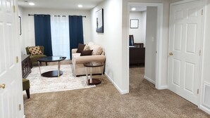 A living room with a beige sofa, a small round table, and a dark-colored armchair. There is a large window with blue curtains, a door leading to another room, and a framed picture on the wall. - Stunning 2400 Sqft. 4 BR, 3.5 BT in the most convenient location to Quantico (Stafford)