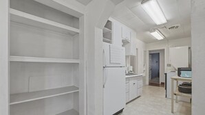 Une cuisine avec des meubles blancs, un réfrigérateur et un évier. On y trouve un plan de travail avec un four à micro-ondes et une petite table avec un panier.